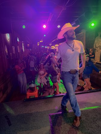Live country singer in a cowboy hat on stage with a microphone as a lively crowd line-dances under colorful neon lights in a rustic bar