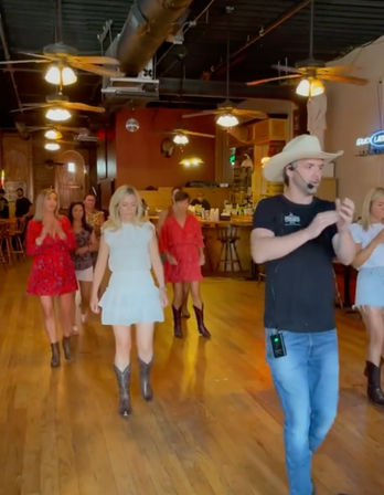 Instructor wearing a cowboy hat leads a line dance in a rustic country bar—women in dresses and cowboy boots on a wooden dance floor under ceiling fans and warm lighting.
