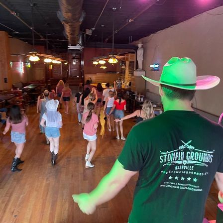 Instructor in a cowboy hat leads a group line dance on a wooden floor in a rustic country bar, dancers in shorts and boots under warm pendant lights.