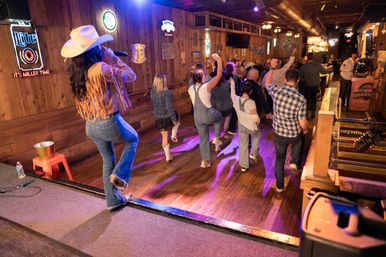 Line dancers in denim and boots on a wooden dancefloor at a lively honky-tonk, led by a cowgirl singer in a fringed jacket and white hat performing from the stage under neon signs