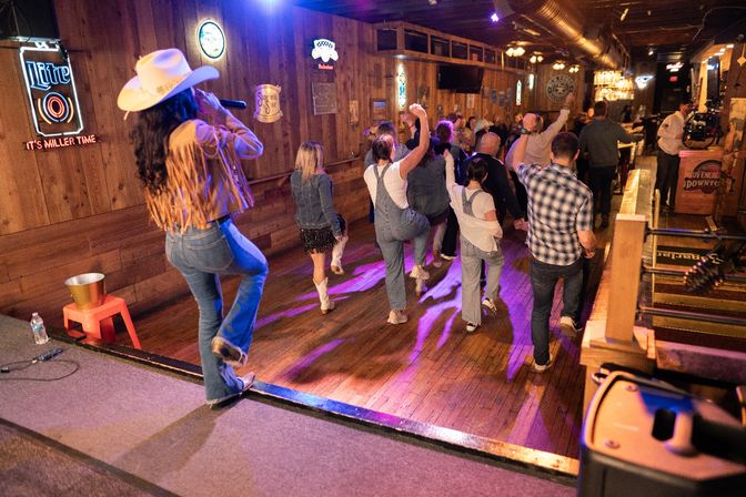 Line dancers in denim and boots on a wooden dancefloor at a lively honky-tonk, led by a cowgirl singer in a fringed jacket and white hat performing from the stage under neon signs