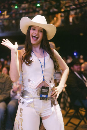 Smiling young woman in a white cowboy hat and sparkling western outfit waving in an arena or rodeo, wearing an artist lanyard and silver belt with a blurred crowd behind her.
