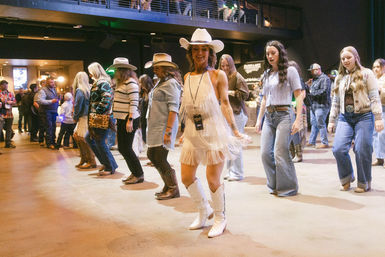 Country line dancing at an indoor dancehall, people wearing cowboy hats, boots and a white fringe dress moving in a row.