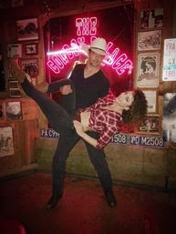 Man in cowboy hat dips smiling woman in a red plaid shirt and cowboy boots in front of a neon sign inside a rustic country bar decorated with license plates and framed photos.
