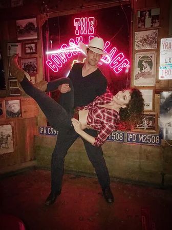 Man in cowboy hat dips smiling woman in a red plaid shirt and cowboy boots in front of a neon sign inside a rustic country bar decorated with license plates and framed photos.