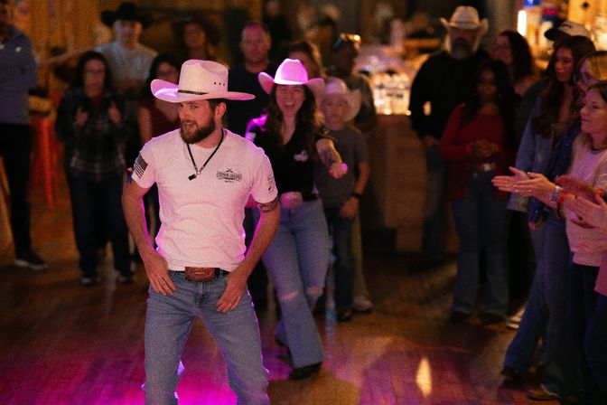 Man in a white cowboy hat and jeans leading a lively country line dance on a wooden bar dance floor, surrounded by a clapping crowd in cowboy hats and warm ambient lighting — country music nightlife scene.
