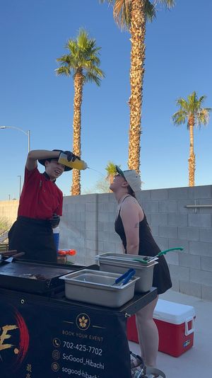 Playful outdoor hibachi chef in red pouring sauce from a squeeze bottle into a guest’s open mouth beside a grill station, prep pans and coolers, with tall palm trees and clear blue desert sky overhead.