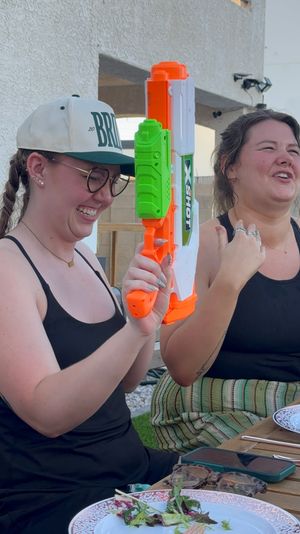 Two friends laughing on a sunny backyard patio, one in a cap and heart-shaped glasses holding a bright orange-and-green water blaster, plates and phones on the wooden table nearby.