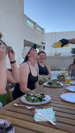 Friends on a sunny backyard patio eating salad — a woman leans back laughing as a gloved hand pours dressing from a yellow squeeze bottle into her open mouth.