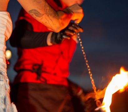 Tattooed arm holding a chained flaming torch in an outdoor nighttime fire-dancing performance, blurred performer in a red shirt and wide-brim hat in the background.