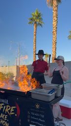Outdoor hibachi chef in a red jacket and wide-brim hat creating dramatic flames as a laughing guest holds a spatula, palm trees and clear blue sky in a sunny backyard setting.