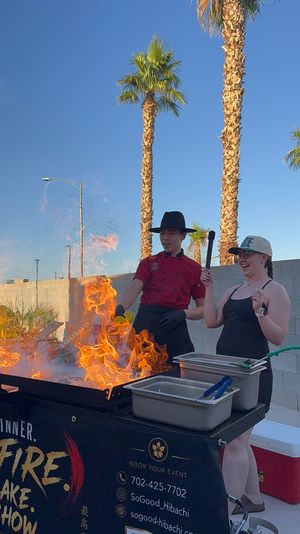 Outdoor hibachi chef in a red jacket and wide-brim hat creating dramatic flames as a laughing guest holds a spatula, palm trees and clear blue sky in a sunny backyard setting.