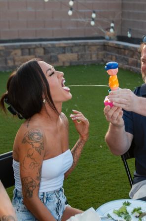 Woman with tattoos at a backyard party on a green lawn playfully leaning back to catch water from a toy squirt held by a friend, stone wall and string lights in the background.