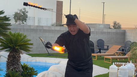 Poolside fire performer twirling a flaming torch at dusk in a backyard patio with lounge chairs, palm plants, and a small swimming pool.