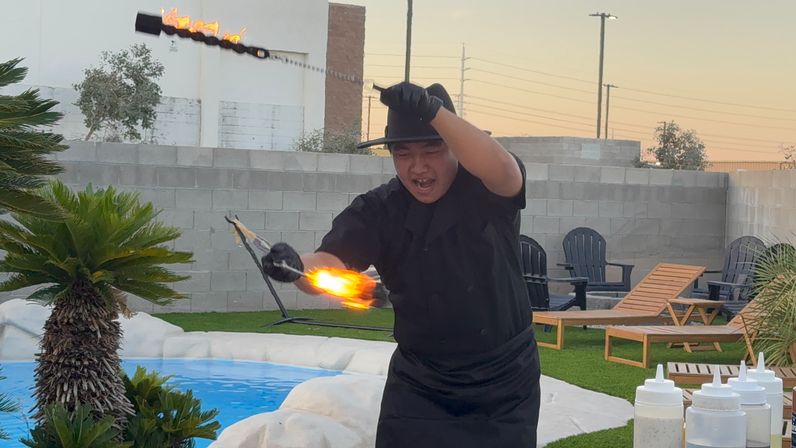 Poolside fire performer twirling a flaming torch at dusk in a backyard patio with lounge chairs, palm plants, and a small swimming pool.