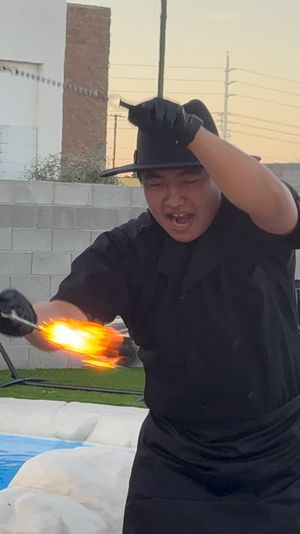 Poolside fire performer twirling a flaming torch at dusk in a backyard patio with lounge chairs, palm plants, and a small swimming pool.