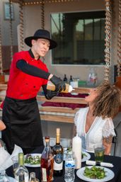 Outdoor patio dinner scene: server in a red chef jacket and black hat playfully pouring dressing from a squeeze bottle into a guest’s open mouth while a table with salads, wine bottles and water sits in front of them.