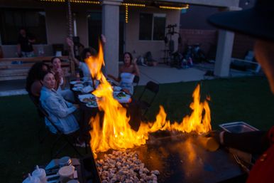 Backyard patio dinner with friends seated at a folding table as a cook flambés food on a sizzling outdoor griddle, tall orange flames illuminating the evening lawn.