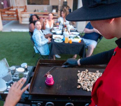 Backyard outdoor dinner party with a cook in a red jacket and black hat flipping a cup on a flat-top griddle beside a pile of sautéing mushrooms, guests seated at a long table in the background.