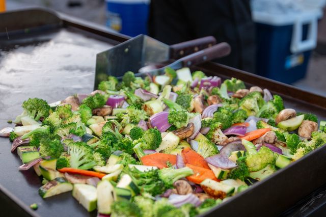 Sizzling colorful mixed vegetables—broccoli, zucchini, mushrooms, red onion and carrots—cooking on a large flat-top griddle with metal spatulas