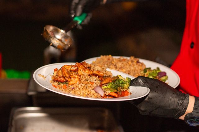 Server hands in black gloves serving white plates of fried rice topped with glazed grilled chicken, broccoli, carrots and onions in a restaurant kitchen.