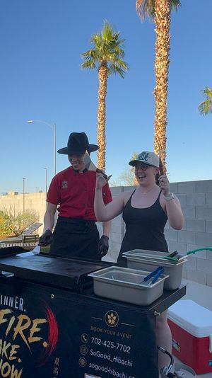 Two people at an outdoor hibachi grill — a chef in a red jacket and black hat and a smiling woman in a baseball cap holding spatulas, standing by prep pans with palm trees and clear blue sky overhead.