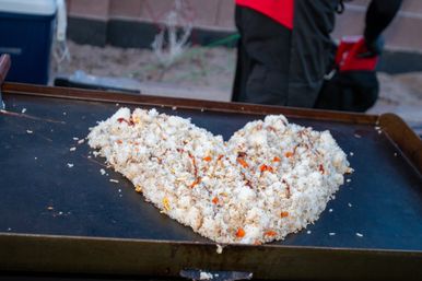 Close-up of seasoned fried rice with diced carrots and scrambled egg cooking on a large outdoor griddle, blurred vendor in a red apron in the background