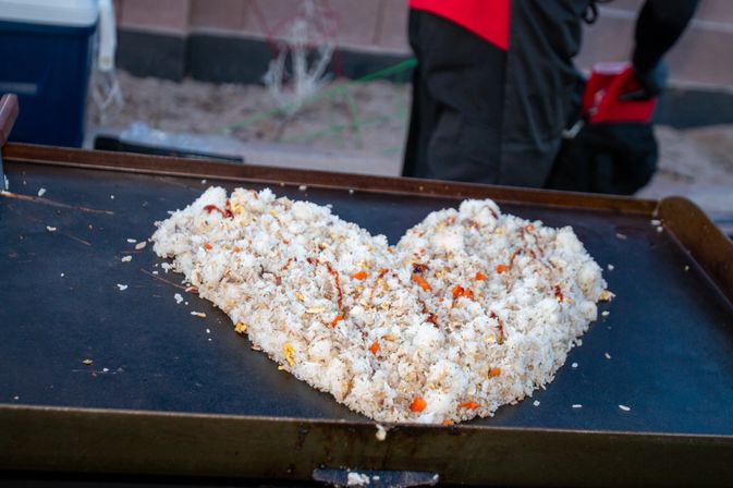 Close-up of seasoned fried rice with diced carrots and scrambled egg cooking on a large outdoor griddle, blurred vendor in a red apron in the background