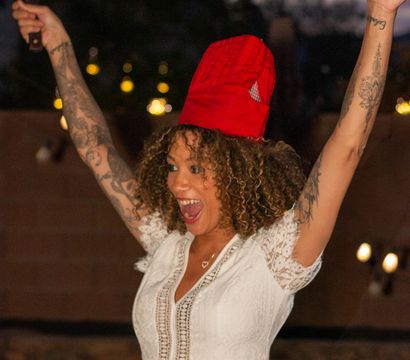 Person in white lace dress and red chef hat cheering with arms raised while holding a spatula at an outdoor backyard evening party, curly hair and visible tattoos