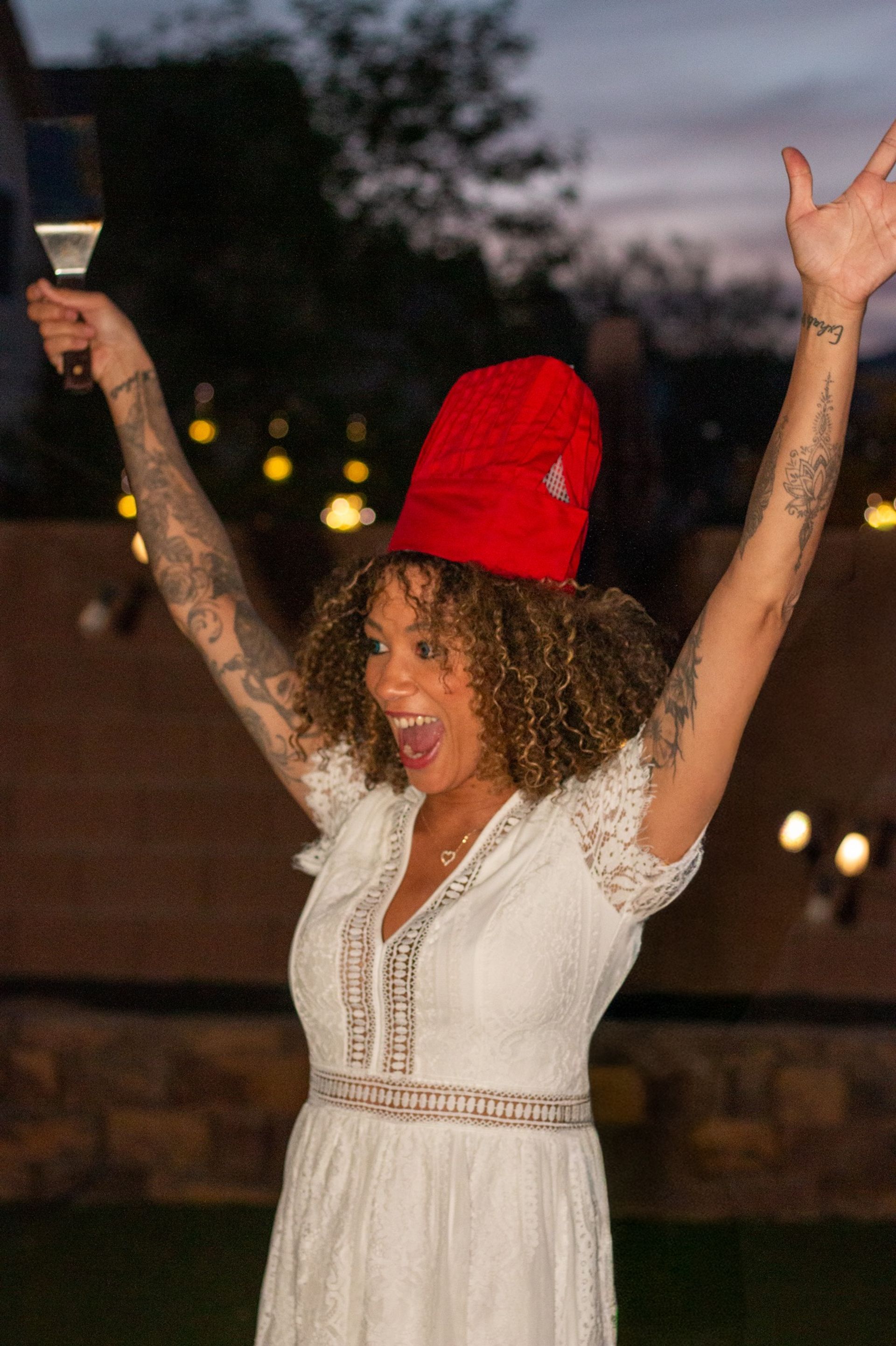 Person in white lace dress and red chef hat cheering with arms raised while holding a spatula at an outdoor backyard evening party, curly hair and visible tattoos