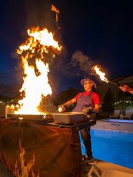 Poolside chef at night flambéing on an outdoor griddle — giant column of fire, assistant with torch, hat-and-sunglasses cook beside a backyard pool with string lights.