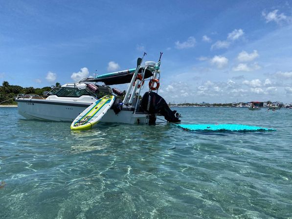 White motorboat anchored in shallow clear turquoise water with a striped stand-up paddleboard leaning on the stern, a turquoise floating mat by the outboard motor, and a sunny blue sky with distant boats and shoreline.