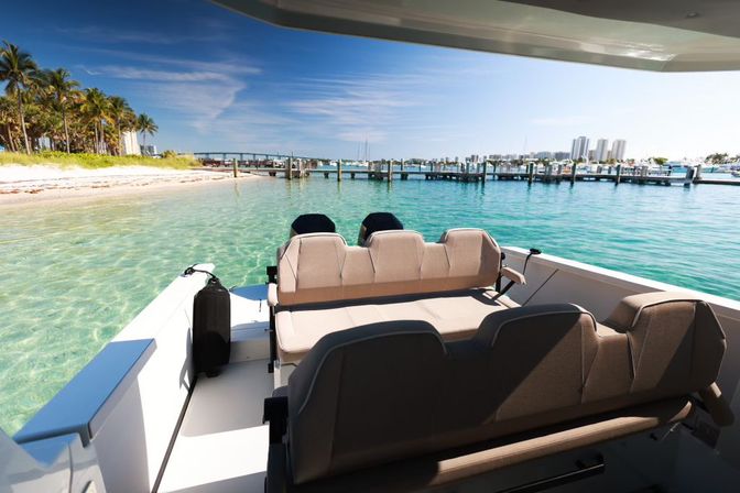 Sunlit boat seating overlooking crystal-clear turquoise water, a sandy palm-lined shore, marina docks and a distant coastal skyline under a bright blue sky.