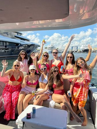 Group of smiling women in colorful swimwear waving and holding drinks on a sunny boat party at a marina with luxury yachts and blue skies in the background.