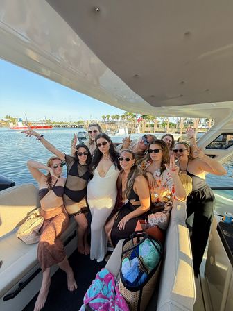 Smiling group of women posing on a yacht deck at a sunny coastal marina with palm trees, docks, beach towels and sunglasses.