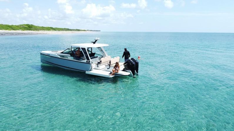 Motorboat anchored in crystal-clear turquoise water near a sandy coastline, three people relaxing on the swim platform under a sunny blue sky