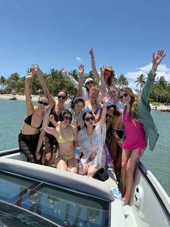 Group of women in swimsuits cheering and waving on a motorboat near a palm‑lined tropical beach with clear turquoise water and a bright blue sky — sun‑filled boat party scene.