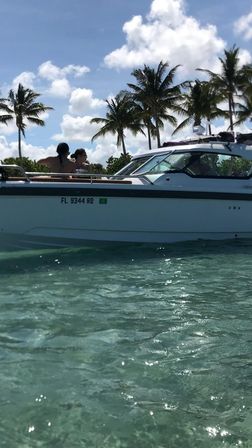 White motorboat anchored in clear turquoise Florida waters, two passengers relaxing on deck beneath palm trees and a bright blue sky with fluffy clouds.