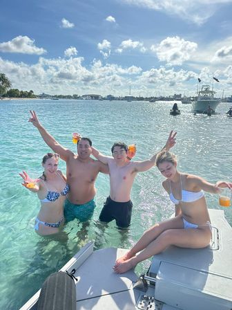 Cheerful group of four in swimsuits holding drinks in clear turquoise water beside a boat under a sunny tropical sky with anchored boats and a palm-fringed shore in the background
