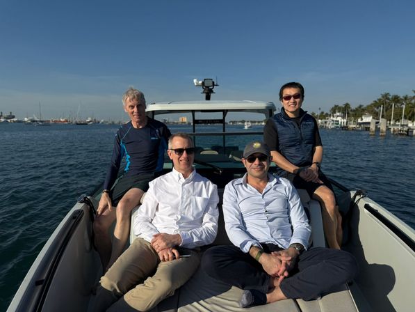 Four men relaxing and smiling on the bow of a motorboat in a sunny coastal marina, calm blue water with boats and palm trees along the shoreline.