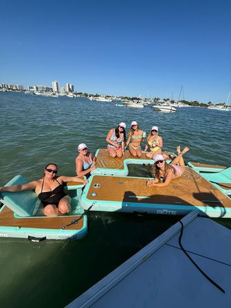 Six women in swimsuits and caps lounging and smiling on turquoise floating boards beside a boat, sipping drinks on a sunny day in a busy marina with yachts and high-rise condos on the shoreline