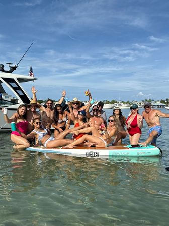 Boat party at a shallow coastal sandbar: a large group of adults in swimsuits posing on a giant paddleboard and beside a yacht, raising drinks and smiling under a sunny blue sky with anchored boats in the background.