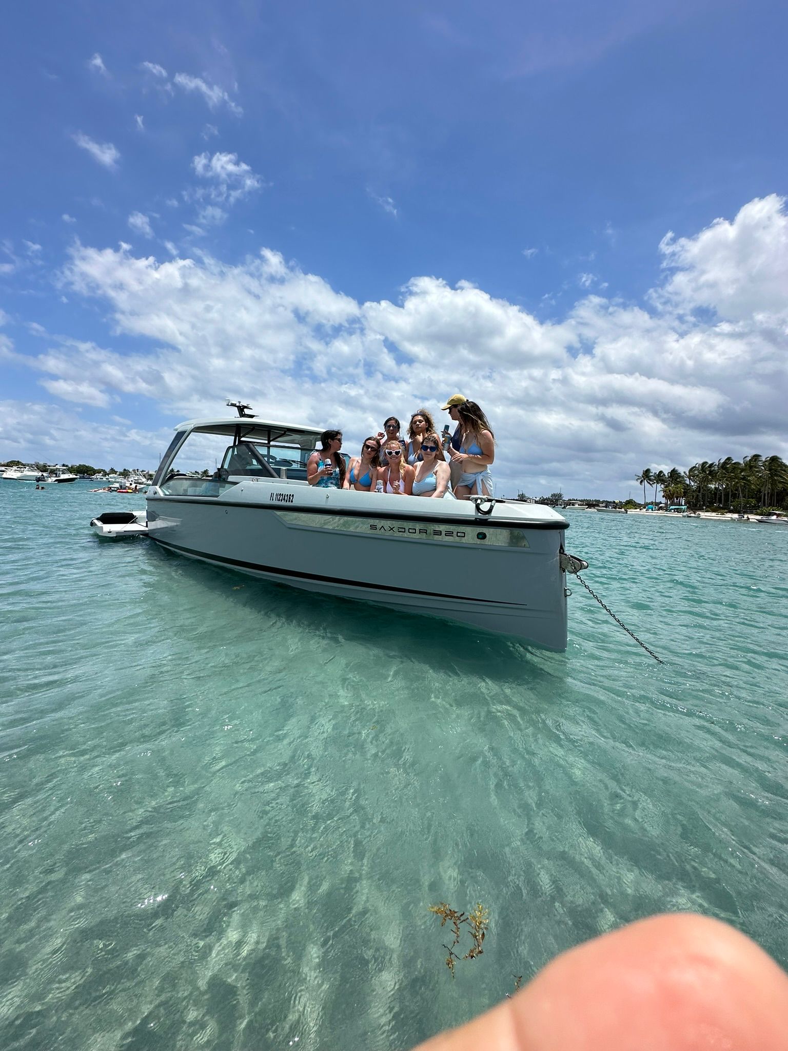 Group of friends enjoying a white motorboat anchored in clear turquoise tropical water near a palm-lined shore under a bright blue sky with puffy clouds.