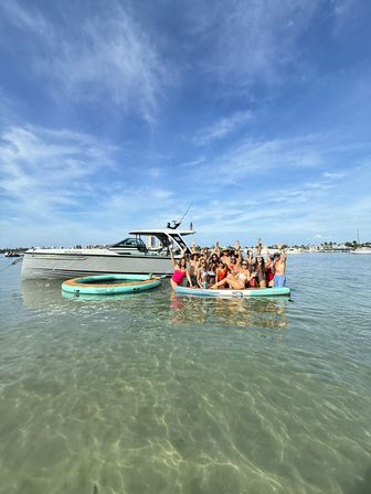 Group of people celebrating on a large paddleboard next to an anchored motorboat at a shallow sandbar with crystal-clear water and a bright blue sky