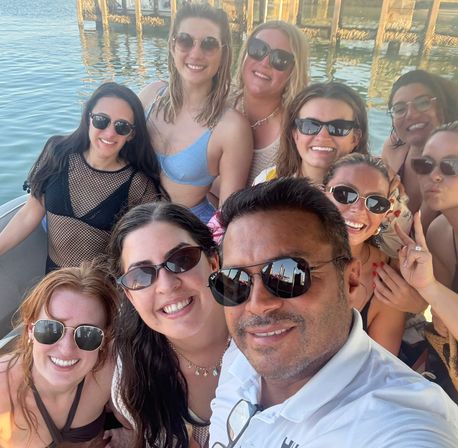 Boat selfie of a smiling group of friends in sunglasses and swimwear on a sunny waterfront near a wooden pier