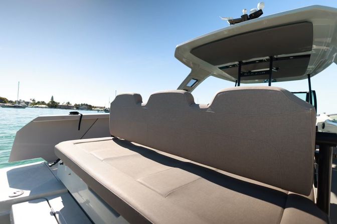 Rear cushioned bench seat on a motorboat with a T-top, overlooking turquoise water and a sunny marina shoreline