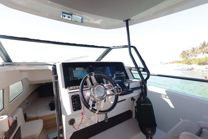 Modern boat helm with polished stainless-steel steering wheel, dual touchscreen navigation displays and throttle controls, cabin entrance to port, fender hanging at the side, and turquoise coastal waters with a palm-lined shoreline visible through the windshield.