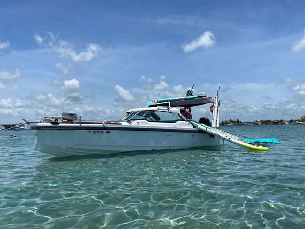 White cabin boat anchored in crystal-clear turquoise water with a paddleboard attached, sunny sky and distant shoreline.