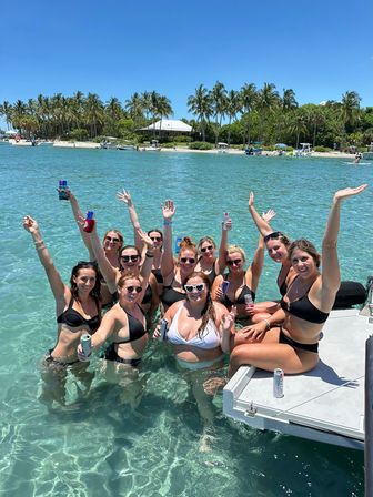 Group of women in bikinis cheering with canned drinks while waist-deep in clear turquoise water beside a boat dock, palm trees and a sunny tropical beach in the background