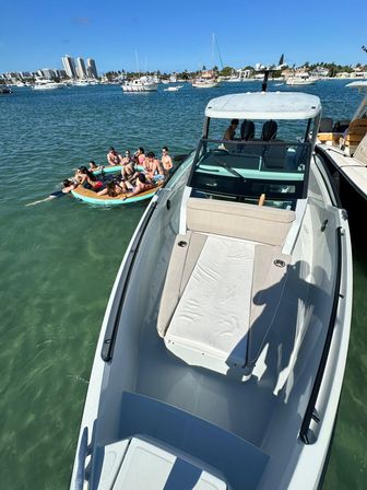 Sunlit coastal scene: overhead view of a white motorboat's bow with cushioned sunpad and a group of people lounging on a round inflatable raft in clear turquoise bay, surrounded by anchored yachts and a palm-lined shoreline with mid-rise buildings.